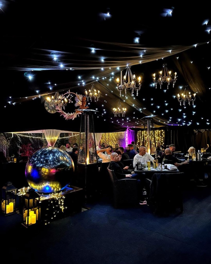 An elegantly decorated indoor venue featuring black drapery and sparkling fairy lights. Chandeliers hang from the ceiling, while a large, reflective sphere and candles create a warm ambience. Guests are seated at tables, engaging in conversation.